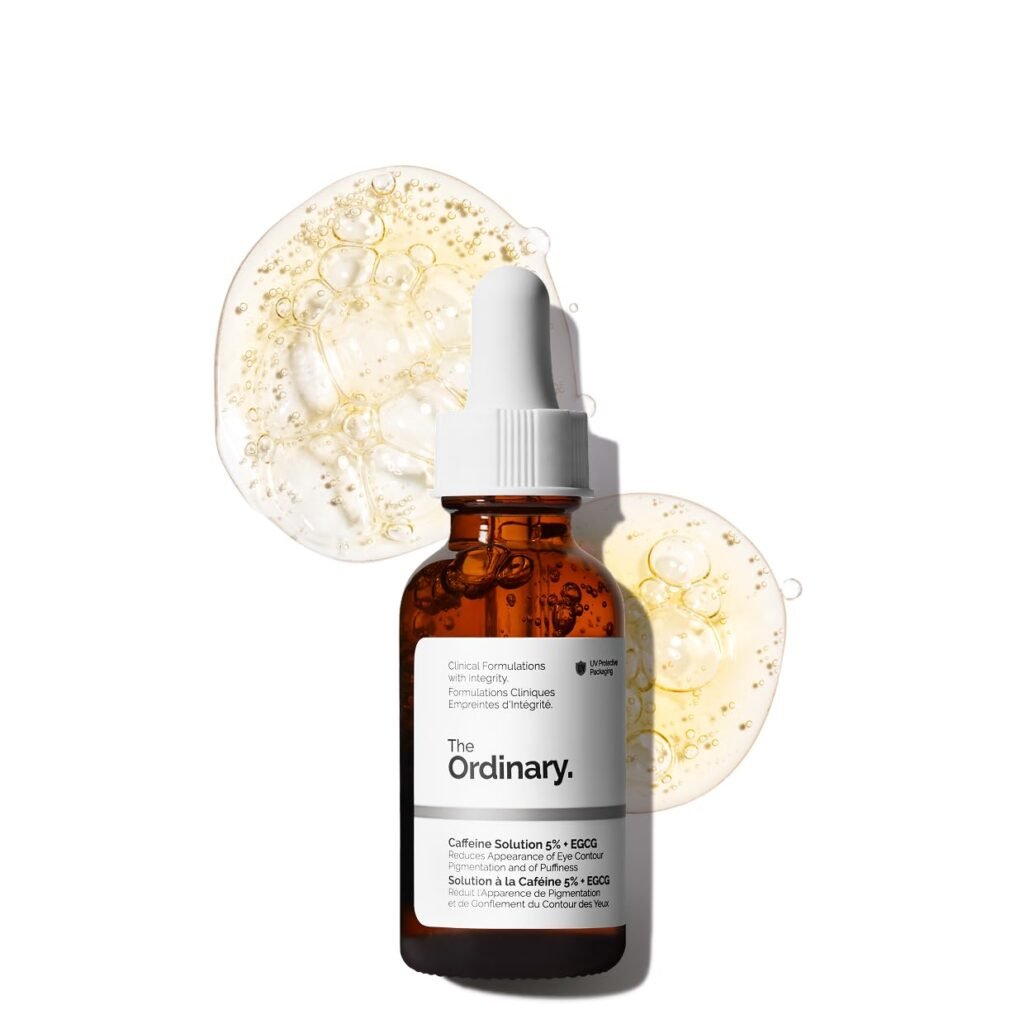 The Deep Dive into What Causes Dark Under Eye Circles? A brown glass dropper bottle of The Ordinary Caffeine Solution 5% + EGCG is shown in front of two translucent drops of serum with bubbles, highlighting its targeted formula for addressing what causes dark under eye circles, on a white background.