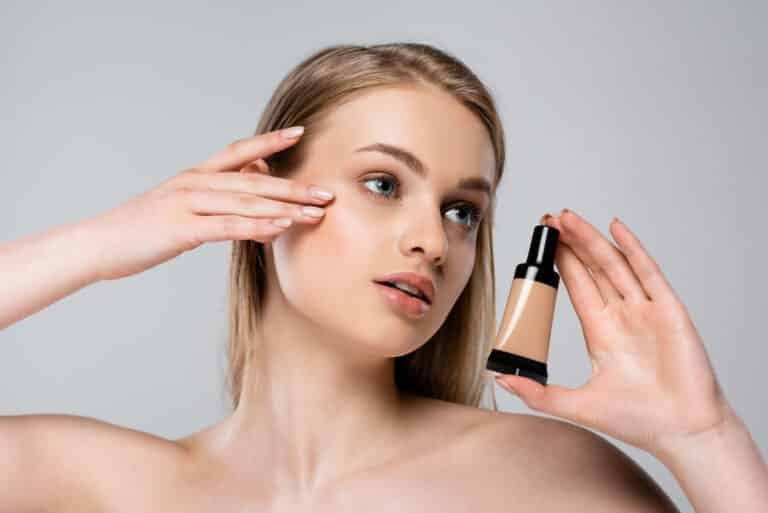 pretty young woman with bare shoulders holding tube with makeup foundation isolated on grey